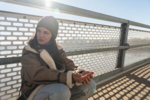 A woman wearing a beanie, brown coat with shearling lining, and jeans sits outside on a sunlit bridge, with her hands clasped and a thoughtful expression on her face.