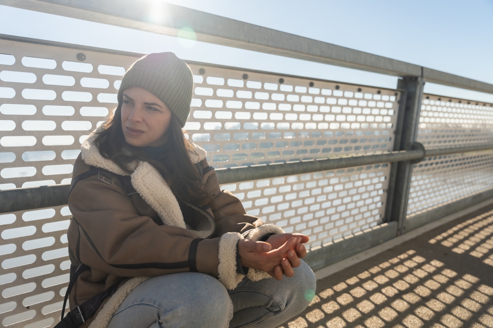 A woman wearing a beanie, brown coat with shearling lining, and jeans sits outside on a sunlit bridge, with her hands clasped and a thoughtful expression on her face.