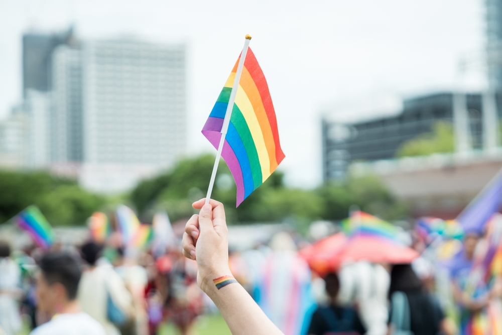 A hand holding a small rainbow pride flag, with a rainbow tattoo on the wrist, at an outdoor event with a crowd and city buildings blurred in the background.