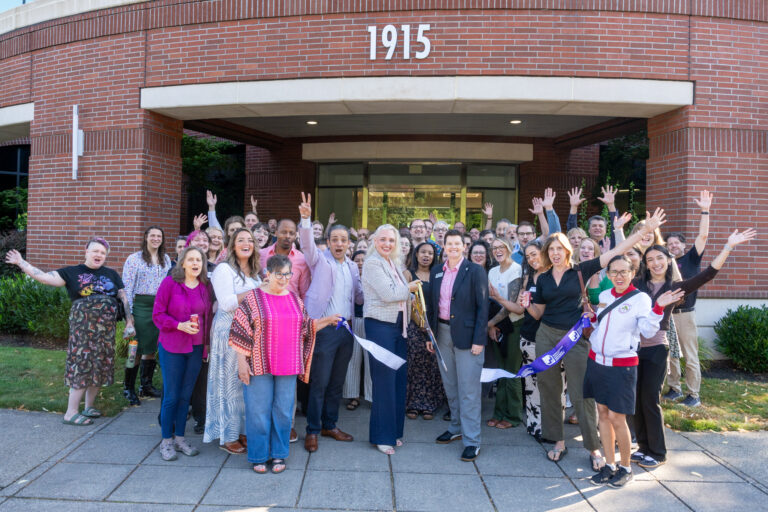 A large, diverse group of people stand outside a brick building labeled “1915,” smiling and raising their arms as a ribbon is cut at the entrance, celebrating a grand opening or special event.