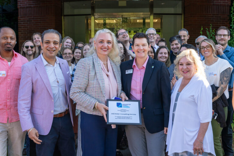 A group of people stand smiling outside a building. Four people in front hold a framed award plaque that reads “2024 Member Counseling Services.” Others in the group stand close together, smiling at the camera.