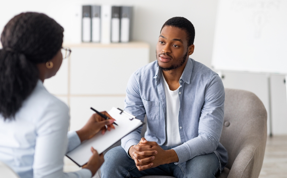 A man sits with his hands clasped, attentively listening to a woman holding a clipboard and pen, suggesting a therapy or counseling session in a modern office setting.