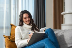 A young woman with long brown hair, wearing a white turtleneck sweater and jeans, sits on a sofa with mustard pillows, smiling as she writes in a notebook. Daylight filters through curtains behind her.