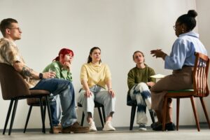 Five people sit in a semi-circle on chairs in a bright room, engaged in a group discussion. One person in blue speaks while the rest listen attentively. The mood appears thoughtful and focused.