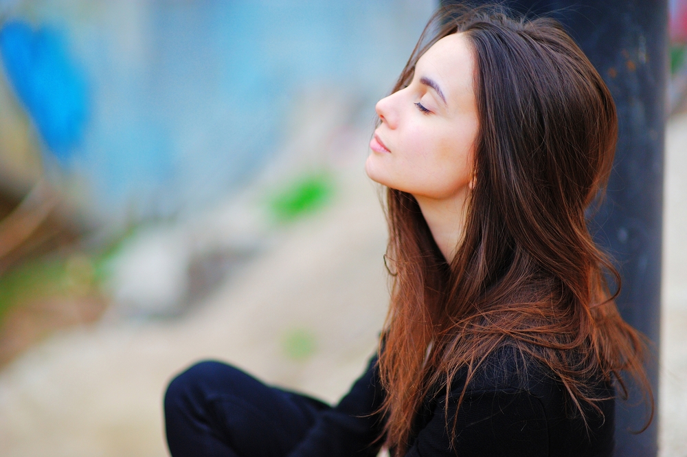 A young woman with long brown hair sits outdoors with her eyes closed, facing upward and appearing serene and relaxed. She wears a black outfit and the background is softly blurred.
